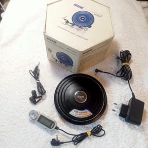 Puede incluir: Un reproductor de audio y vídeo portátil negro con una caja a juego, auriculares y un cargador. El reproductor tiene un diseño circular con el texto "FM-тюнер" y "VCD-MP3-CD-DA". La caja tiene el texto "PV430T".