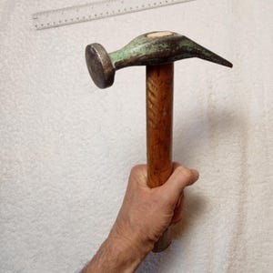 Può includere: Un martello vintage con manico in legno e testa in metallo verde. Il martello ha una testa arrotondata e un'unghia leggermente curva.