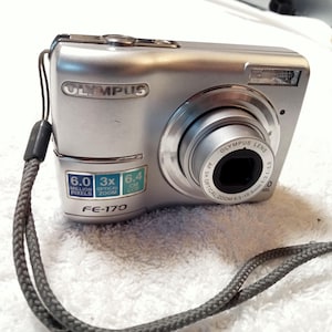 May include: Silver Olympus FE-170 digital camera with a lens, flash, and wrist strap. Features include 6.0 million pixels, 3x optical zoom, and a 6.4 cm LCD screen. The camera has the Olympus logo.
