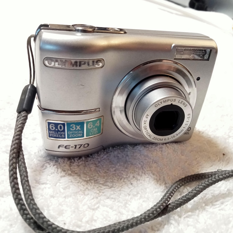 May include: Silver Olympus FE-170 digital camera with a lens, flash, and wrist strap. Features include 6.0 million pixels, 3x optical zoom, and a 6.4 cm LCD screen. The camera has the Olympus logo.