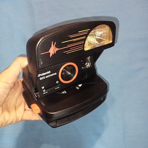 May include: Black Polaroid 600 extreme instant camera with a red and orange stripe design. The camera has a built-in flash and a lens cover.