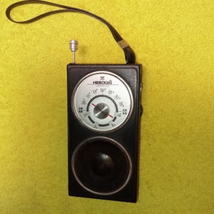 May include: A vintage black portable radio with a silver dial and speaker. The dial features the text "HEBCKИЙ" and a tuning scale. A black leather strap is attached to the top. The radio is set against a yellow background.