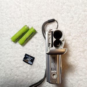 May include: A silver digital camera with an open battery compartment, two green Esperanza batteries, and an Olympus XD M512MB memory card. The camera has a black wrist strap attached. The items are on a white, textured surface.