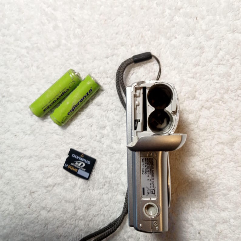 May include: A silver digital camera with an open battery compartment, two green Esperanza batteries, and an Olympus XD M512MB memory card. The camera has a black wrist strap attached. The items are on a white, textured surface.