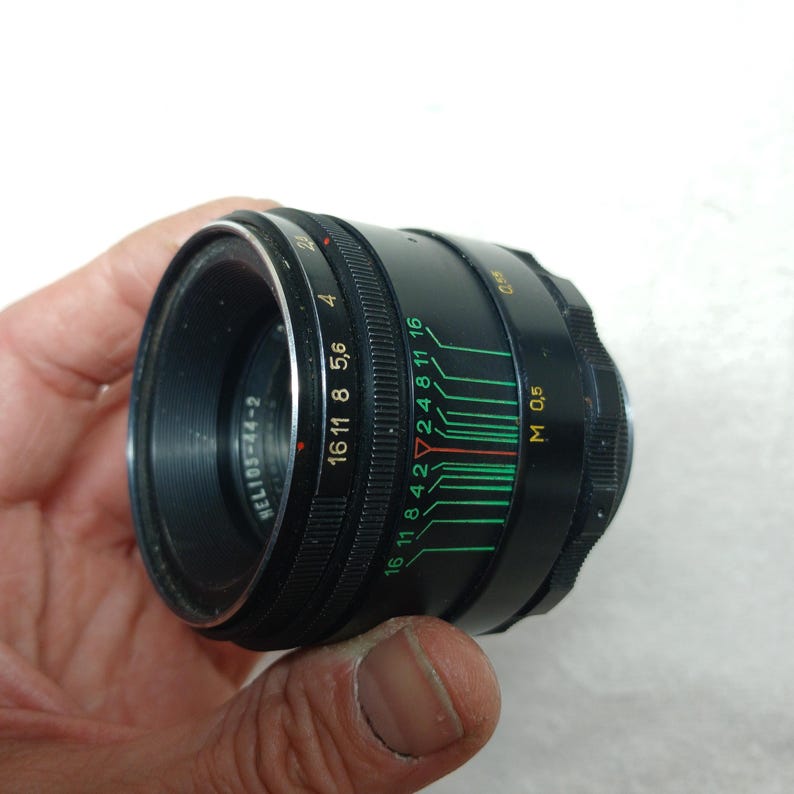 Peut inclure: Un objectif d'appareil photo noir Helios-44-2 avec des marquages verts et blancs. L'objectif a une bague de mise au point et des r&eacute;glages d'ouverture de 2 &agrave; 16. L'objectif est tenu dans une main.