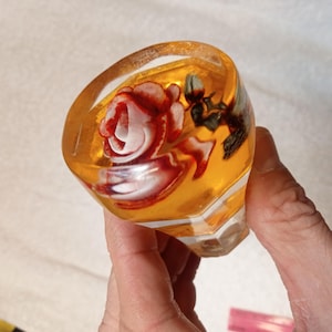 Op de afbeelding: Een decoratieve flesstopper met een rozenontwerp. De stopper is gemaakt van een helder, amberkleurig materiaal, met een rode en witte roos in het midden. De stopper heeft een facetgeslepen basis.