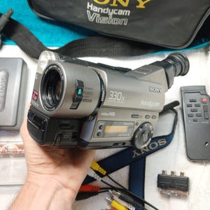 May include: A silver and black Sony Handycam video Hi8 camcorder with 330x digital zoom, featuring a lens with 16x optical zoom, a small LCD display, a power switch, and various control buttons. The device includes a remote control, cables, and a bag.
