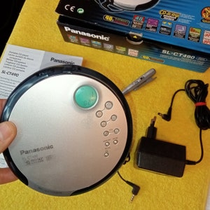 Könnte beinhalten: Ein silberner und schwarzer Panasonic SL-CT490 tragbarer CD-Player mit digitalem Display und Tasten für Lautstärke, Speicher, Modus und Wiederholung. Der Player ist mit einem schwarzen Netzteil und Kopfhörern verbunden.