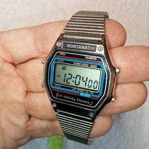 May include: A silver Montana digital watch with a rectangular face. The watch face displays the time as 12:04:00 PM. The watch has a metal strap and features buttons for light, start/stop, date, mode, and select functions. The watch also has a 16-Melody Chrono function.