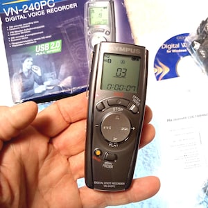 Op de afbeelding: Een donkergrijze Olympus VN-240PC digitale voicerecorder met een klein LCD-scherm dat cijfers en tijd weergeeft. De recorder heeft verschillende knoppen voor afspelen, stoppen en menufuncties. De doos op de achtergrond heeft de tekst "USB 2.0 FULL SPEED".