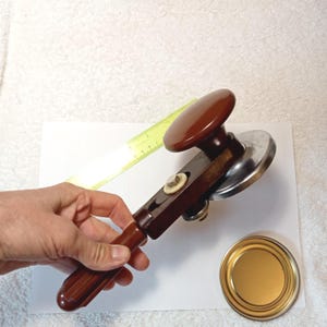 以下が含まれることがあります： 茶色と銀色のヴィンテージ缶切り。茶色のハンドルとノブが付いています。 金色の缶の蓋の隣に缶切りが表示されています。 黄色の定規が背景にあります。