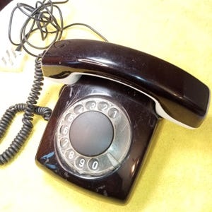 May include: A vintage black rotary dial telephone with a glossy finish. The phone features a circular dial with numbers and a matching handset. The coiled cord and power cord are visible, suggesting it is a landline phone.