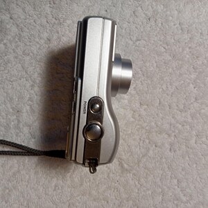 May include: Silver digital camera with a lens, power button, and wrist strap. The camera has a sleek, compact design. The word "POWER" is printed on the side of the camera.