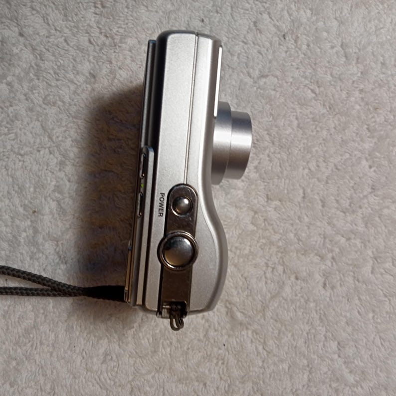 May include: Silver digital camera with a lens, power button, and wrist strap. The camera has a sleek, compact design. The word "POWER" is printed on the side of the camera.