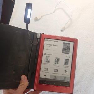 May include: A red Sony e-reader with a black protective cover and an attached flexible LED reading light. The screen displays a menu with book titles including The Da Vinci Code and various icons for books, periodicals, collections, and notes.