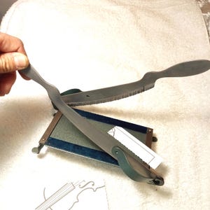 May include: A vintage metal violin bridge cutter with a serrated blade. The cutter has a dark metal finish and a wooden base. The handle is held by a hand. A small piece of paper is attached to the base.