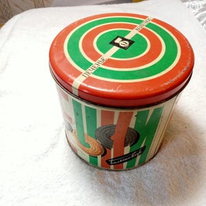 Op de afbeelding: Vintage ronde blikken doos met een rood, groen en wit doelontwerp op het deksel. De zijkant van de doos heeft verticale groene en witte strepen en illustraties van koekjes. Tekst in het Russisch is zichtbaar.