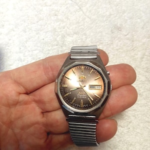 May include: A vintage Orient AAA Crystal wristwatch with a brown and gold dial. The watch has a silver-tone metal band and case. The day and date are displayed at the 3 o'clock position. The watch is held in a hand.