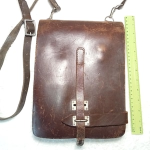 May include: A brown leather messenger bag with a shoulder strap and metal hardware. The bag has a rectangular shape with a flap closure and a buckle. A ruler is placed next to the bag. The bag measures approximately 10 inches in height.