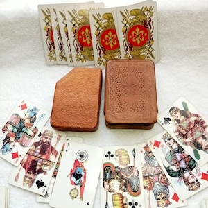 May include: A vintage deck of playing cards with ornate illustrations and a brown leather card case. The cards feature detailed artwork of historical figures and symbols. The card case is a rectangular shape with a textured surface.