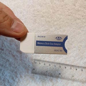 以下が含まれることがあります： シルバーと白のソニーメモリースティックデュオアダプター。「SONY」と「Memory Stick Duo Adaptor」の文字が印刷されています。モデル番号「MSAC-M2」も表示されています。