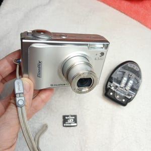 May include: A silver Fujifilm FinePix digital camera with a Fujinon zoom lens, a gray wrist strap, a black battery charger, and a small black xD-Picture Card memory card. The camera body features the FinePix logo and 6.3 Mega Pixels text.