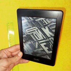 Könnte beinhalten: Ein schwarzer Kindle E-Reader mit einem grau-weißen Muster auf dem Bildschirm. Der Bildschirm zeigt unten das Wort "kindle" an.
