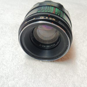 Peut inclure: Un objectif d'appareil photo Helios-44-2 noir avec des marquages verts, rouges et blancs. L'objectif a une bague argent&eacute;e et un corps noir avec des indicateurs de mesure. L'objectif est inclin&eacute; vers le spectateur, mettant en valeur ses &eacute;l&eacute;ments en verre.