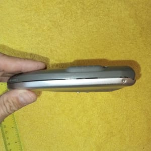 May include: A silver and gray handheld electronic device with a small button on the top. The device is held in a hand.