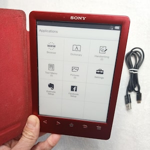 以下が含まれることがあります： 赤いSony製電子書籍リーダーと、お揃いの赤い保護カバー。画面には「Applications」メニューが表示されており、Browser、Dictionary、Handwriting、Text Memo、Pictures、Settings、Evernote Setup、Facebook Setupのアイコンが並んでいます。黒いUSBケーブルが付属しています。
