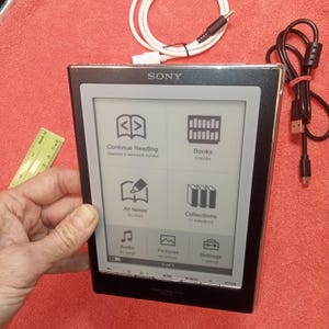 Puede incluir: Lector de libros electrónicos Sony negro con borde plateado, que muestra un menú con opciones para leer, libros, notas y configuraciones. Está conectado a un cable USB blanco y negro. Una regla es visible en el fondo.