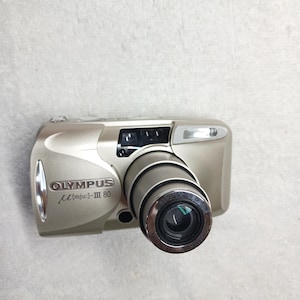 May include: A champagne-colored Olympus Mju-III 80 compact film camera featuring a retractable zoom lens. The front body displays the Olympus brand name and model text, a built-in flash unit, and a viewfinder window positioned above the lens barrel.