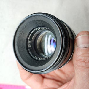 Peut inclure: Un objectif d'appareil photo noir avec le num&eacute;ro "7460677" imprim&eacute; sur le bord. L'objectif a une bague de mise au point stri&eacute;e et un &eacute;l&eacute;ment en verre transparent. L'objectif est tenu dans une main.