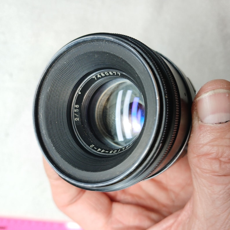 Peut inclure: Un objectif d'appareil photo noir avec le num&eacute;ro "7460677" imprim&eacute; sur le bord. L'objectif a une bague de mise au point stri&eacute;e et un &eacute;l&eacute;ment en verre transparent. L'objectif est tenu dans une main.