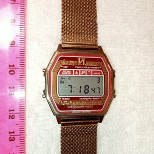 May include: A vintage digital watch with a gold-tone mesh band and a red face. The watch displays the time as 7:18:47 and includes Cyrillic text. The watch face has a rectangular shape with an octagonal frame. The watch is approximately 1.5 inches wide.