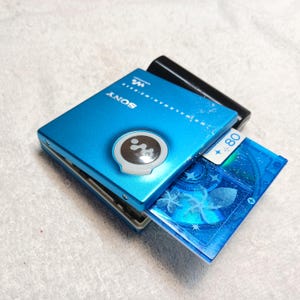 May include: A bright blue Sony Walkman MD player with a circular silver logo and the text SONY and MD WALKMAN MZ-E510. A blue MiniDisc with a floral and star pattern and the number 80 is partially inserted into the side of the portable device.