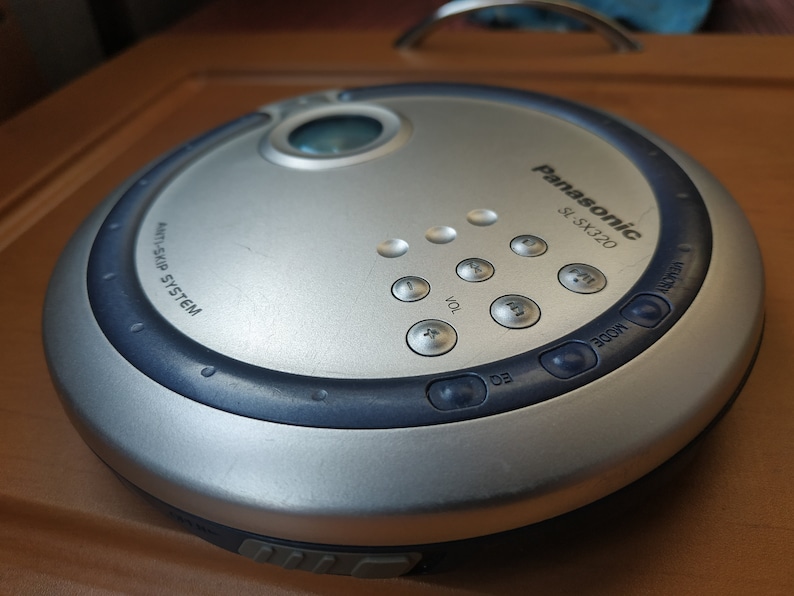 Puede incluir: Un reproductor de CD port&aacute;til Panasonic SL-SX220 plateado con un borde azul y varios botones, que incluyen volumen, modo y salto. El reproductor tiene un sistema anti-salto.