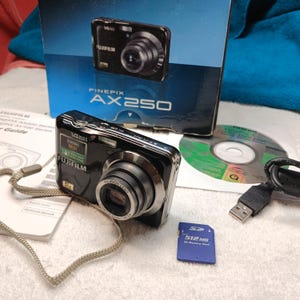 May include: A black Fujifilm FinePix AX250 digital camera with a 5x zoom lens, a wrist strap, a 512MB SD memory card, a USB cable, a software disc, and a user guide, all presented with the original blue and black product packaging box.