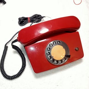 May include: A vintage red rotary dial telephone with a black coiled handset cord. The phone has a circular dial with numbers and a small button on the side. The handset rests on the main body of the phone.