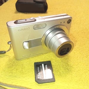 May include: Silver Casio digital camera with a retractable lens. The camera has the words "CASIO" and "OPTICAL ZOOM" printed on it. A black memory card adapter is next to the camera. A black camera case is in the background.