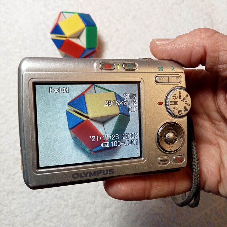 May include: A silver Olympus digital camera displaying a colorful geometric puzzle on its screen. The puzzle is composed of red, yellow, blue, and green triangular pieces. Another puzzle sits above the camera. The screen shows the date and time.