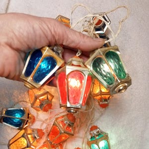 May include: A string of vintage decorative lanterns in various colours, including red, blue, green, and orange. The lanterns have a gold-coloured metal frame and are strung together with a white wire. The lanterns are lit, creating a warm glow.