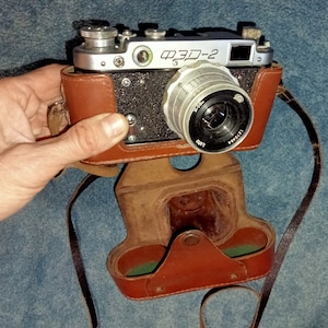 Puede incluir: Cámara ФЭД-2 vintage con funda de cuero marrón. La cámara tiene un cuerpo plateado con una parte frontal texturizada negra y una lente. La funda está abierta, revelando un interior marrón. Una correa negra está adjunta.