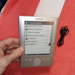 Puede incluir: Lector de libros electrónicos Sony plateado con una pantalla blanca que muestra un menú en ruso. El dispositivo tiene una funda protectora y un cable USB negro. La pantalla muestra opciones como "Continuar leyendo" y "Libros por título".