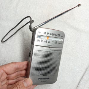 May include: A silver Panasonic RF-P50D portable radio with a black wrist strap and an extended metal antenna. The front features a speaker grille, a tuning dial with FM and AM frequency scales, and the text Panasonic RF-P50D FM-AM 2-BAND RECEIVER.