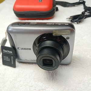 Peut inclure: Appareil photo numérique Canon PowerShot A490 argenté avec objectif et flash noirs. Un étui rouge, une carte mémoire SanDisk et un câble noir sont également visibles. L'appareil photo a un zoom 3,3x.