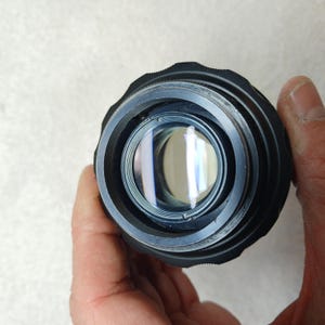 Peut inclure: Gros plan sur un objectif d'appareil photo noir avec un &eacute;l&eacute;ment en verre r&eacute;fl&eacute;chissant. L'objectif a une bague ext&eacute;rieure stri&eacute;e et est tenu par une main. Le verre refl&egrave;te la lumi&egrave;re, cr&eacute;ant un effet circulaire brillant.