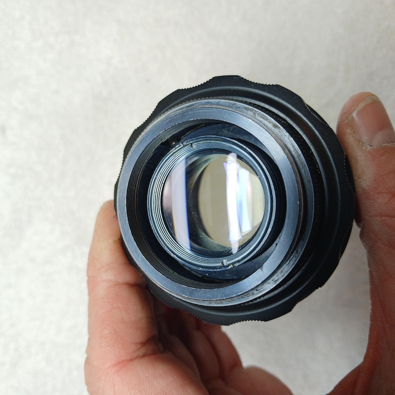 Peut inclure: Gros plan sur un objectif d'appareil photo noir avec un &eacute;l&eacute;ment en verre r&eacute;fl&eacute;chissant. L'objectif a une bague ext&eacute;rieure stri&eacute;e et est tenu par une main. Le verre refl&egrave;te la lumi&egrave;re, cr&eacute;ant un effet circulaire brillant.