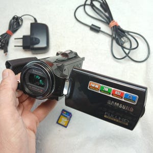 May include: A black Samsung Full HD camcorder with a Schneider Kreuznach lens, 20x optical zoom, and BSI CMOS sensor. The device features a flip-out screen with Full HD 1920x1080 branding, accompanied by a black power adapter and a USB cable.