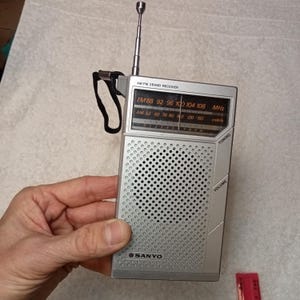 May include: A silver Sanyo AM/FM 2-band receiver radio with a black strap. The radio has a telescoping antenna and a speaker grill. The radio's display shows FM and AM frequencies. The radio has tuning and volume controls.